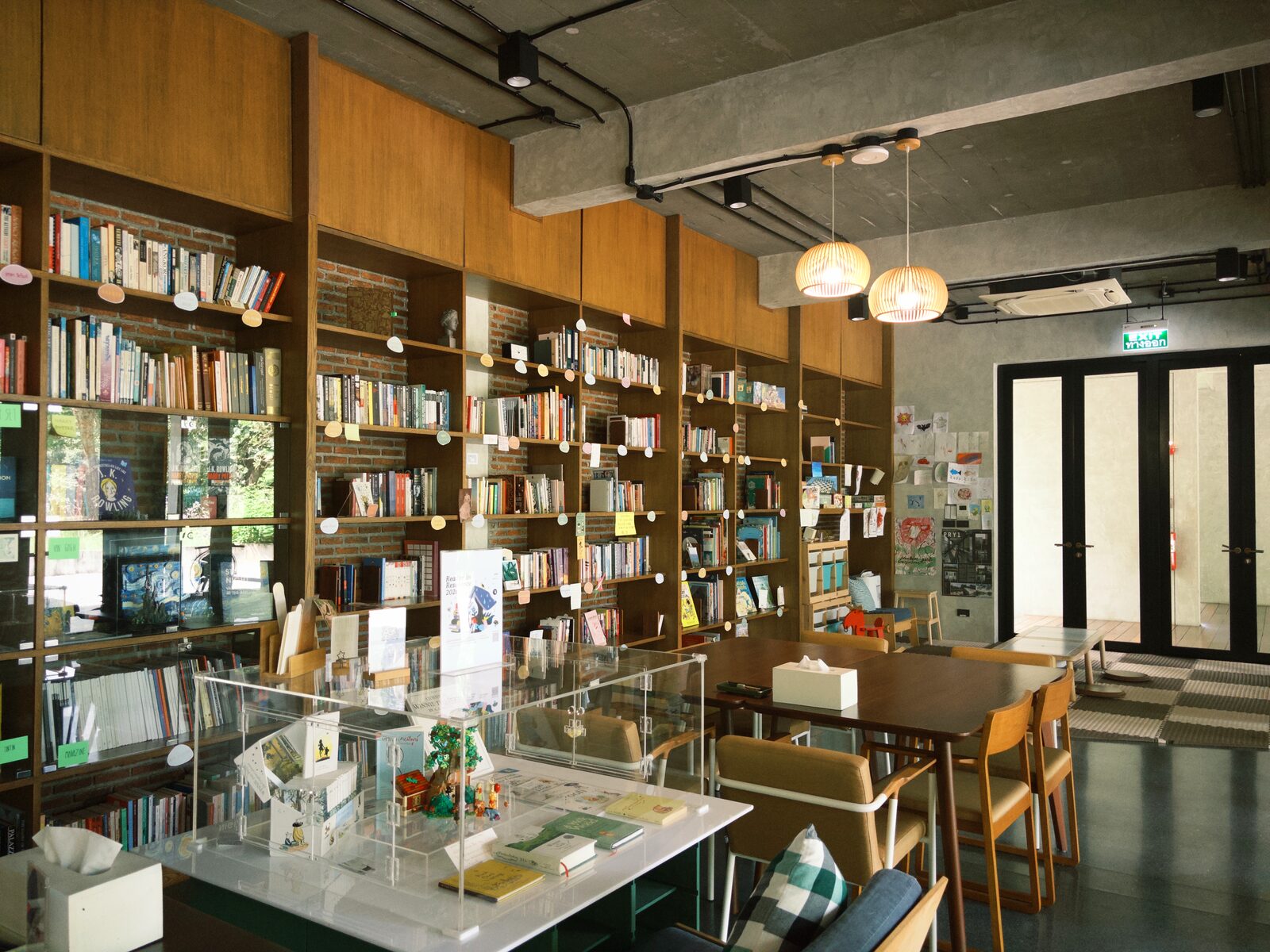 ชั้นหนังสือไม้สูงจรดเพดานคอนกรีตในห้องสมุด bulbul in the woods โคมไฟสานให้แสงอุ่นเหนือโต๊ะไม้และเก้าอี้