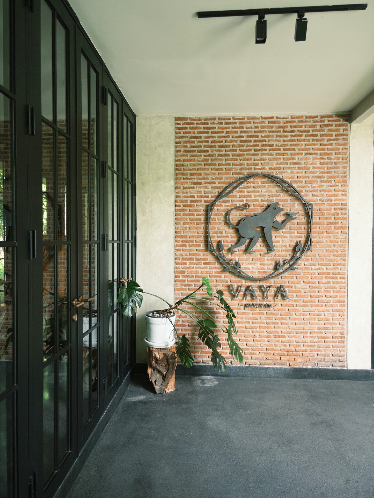 โลโก้โลหะรูปสัตว์ชูใบไม้ในกรอบวงกลมบนผนังอิฐแดง หน้าทางเข้าห้องอาหาร VAYA ประตูกระจกกรอบเหล็กดำ