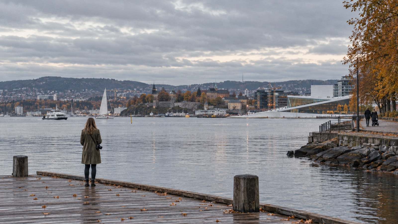 หญิงสะพายกล้องยืนหันหลังบนท่าไม้ริมฟยอร์ดออสโลใต้ฟ้าครึ้ม ใบไม้ร่วงเกลื่อนพื้น ไกลออกไปมีคู่หนึ่งเข็นรถเด็กเดินเลียบทางเดินริมน้ำ