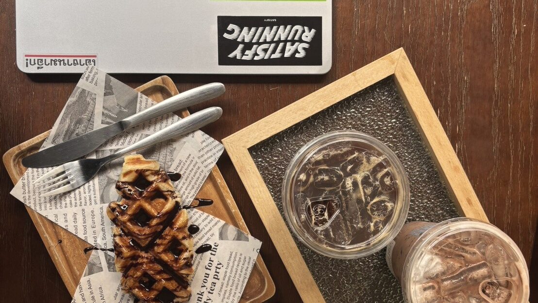 มุมโต๊ะไม้สีเข้มในร้านกาแฟ วาฟเฟิลราดช็อกโกแลตวางบนกระดาษหนังสือพิมพ์ในถาดไม้ ข้าง ๆ มีกาแฟเย็นสองแก้วในถาดไม้กรุกระจก