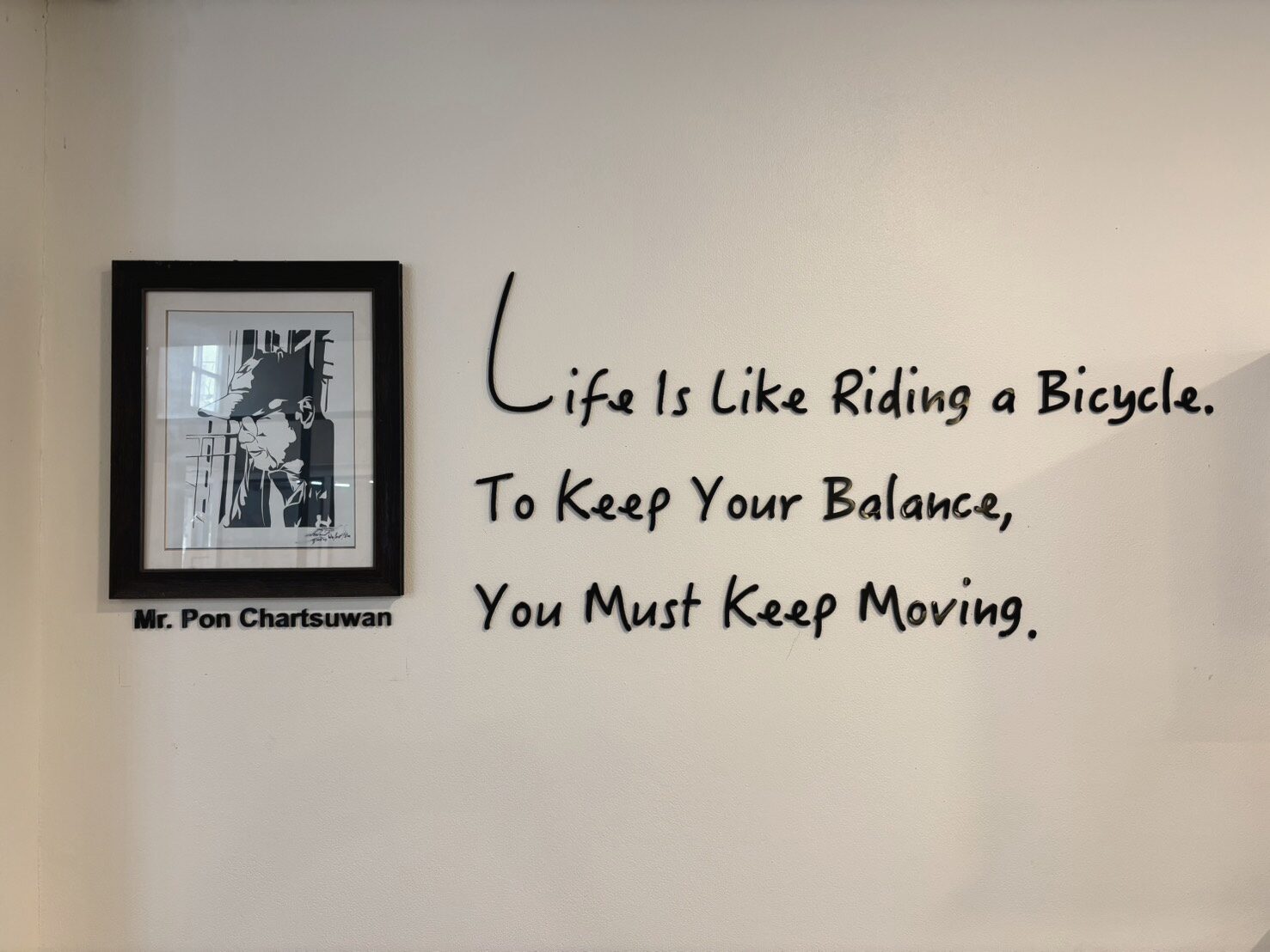 ผนังสีขาวของร้าน Totti Coffee มีประโยคภาษาอังกฤษ “Life is like riding a bicycle. To keep your balance, you must keep moving.” ติดเป็นตัวอักษรสีดำ ข้าง ๆ มีภาพพอร์เทรตขาวดำในกรอบไม้