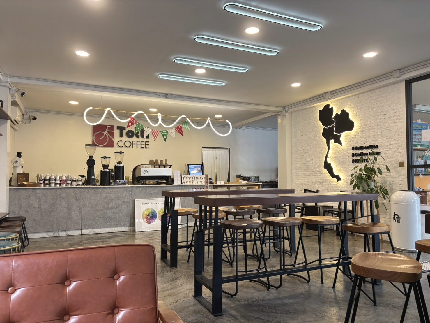 ภายในร้าน Totti Coffee ช่วงบ่าย โต๊ะไม้สูงกับสตูลเรียงอยู่กลางร้าน โซฟาหนังสีน้ำตาลอยู่ด้านหน้า ป้ายนีออนและแผนที่ประเทศไทยติดบนผนังอิฐขาว
